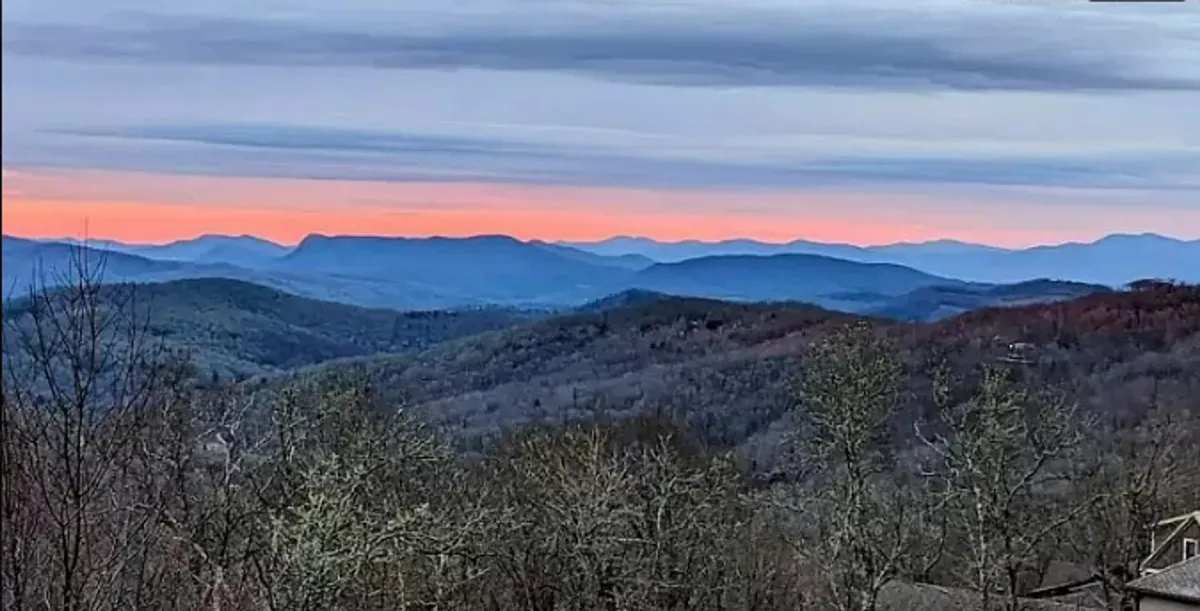 The image showcases a sweeping view of the Blue Ridge mountain range from Sugar Mountain at sunset. The vibrant sky casts warm hues over a densely forested tree line, creating a calm and inviting atmosphere. The vantage point emphasizes the condo’s elevated perch and its unobstructed views of nature.