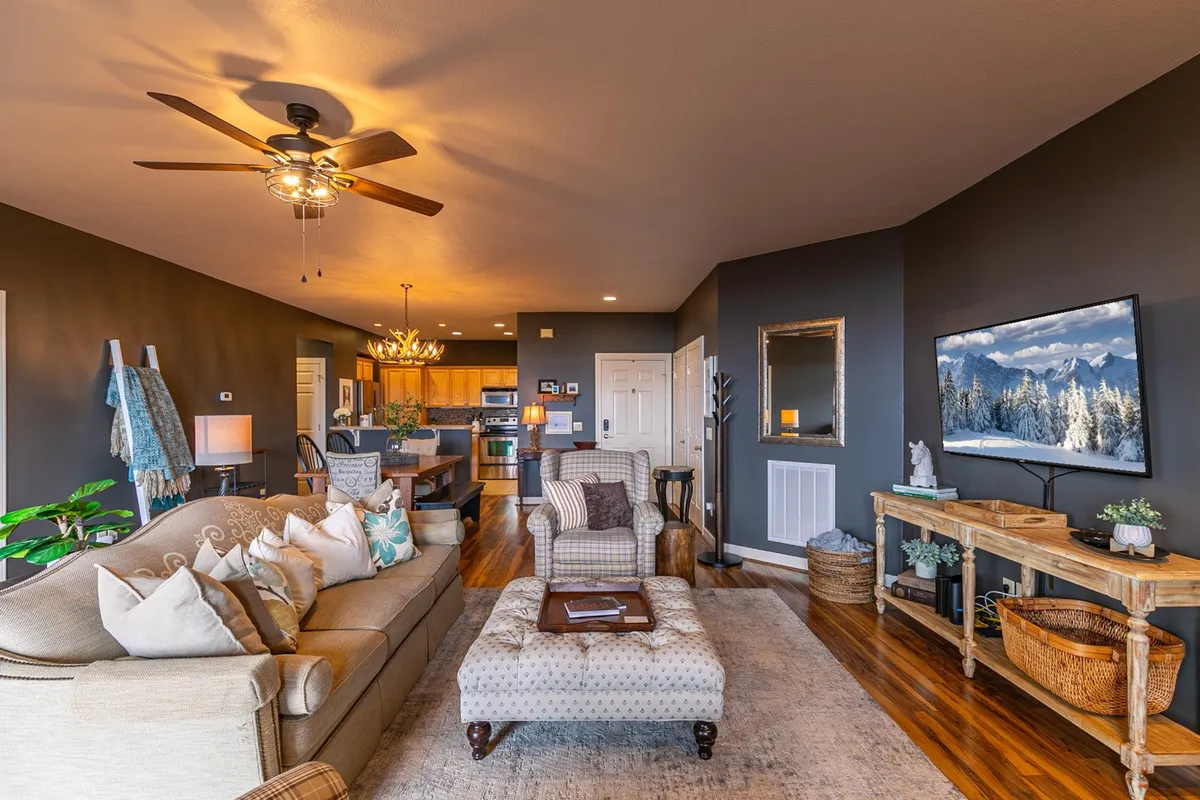 This image highlights a comfortable sofa with decorative pillows, a large TV displaying a snowy mountain scene, and a glimpse of the kitchen area, all under a stylish ceiling fan. The scene is lively yet cozy, suggesting space for family gatherings, movie nights, and relaxed entertaining in an open-concept setting.