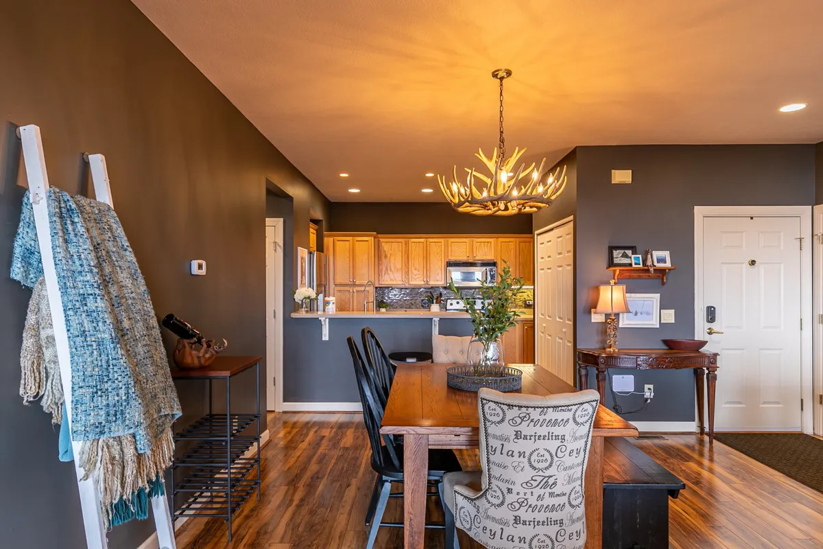 The dining area is depicted with a cozy wooden dining table under a chandelier, seamlessly connected to the kitchen space. Decorative chairs and wall accents add a sense of warmth and personality, evoking the idea of shared meals, game nights, or lingering over wine with mountain glimpses beyond.