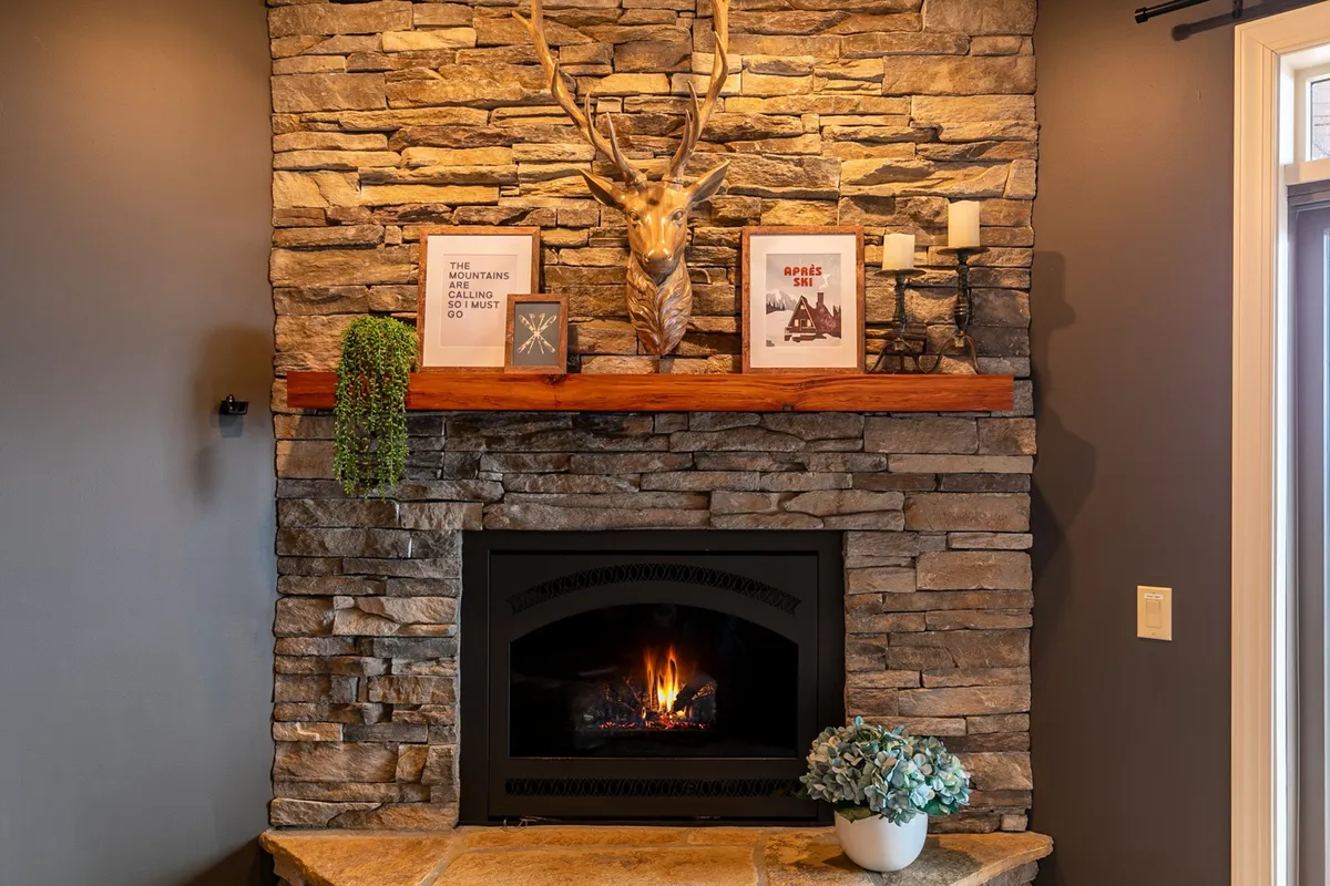 A warm, inviting living room centered around a striking stone fireplace with antler decor above the wooden mantle. A potted plant and decorative frames add a cozy touch, inviting guests to gather, read, or enjoy a hot beverage after a snowy day outdoors.