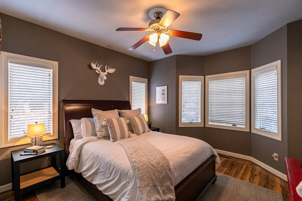 A tranquil bedroom showcasing a queen-size bed with decorative pillows, a ceiling fan, and a window arrangement that lets in soft natural light. The décor includes a whimsical decorative moose head, adding a personalized touch to the space.