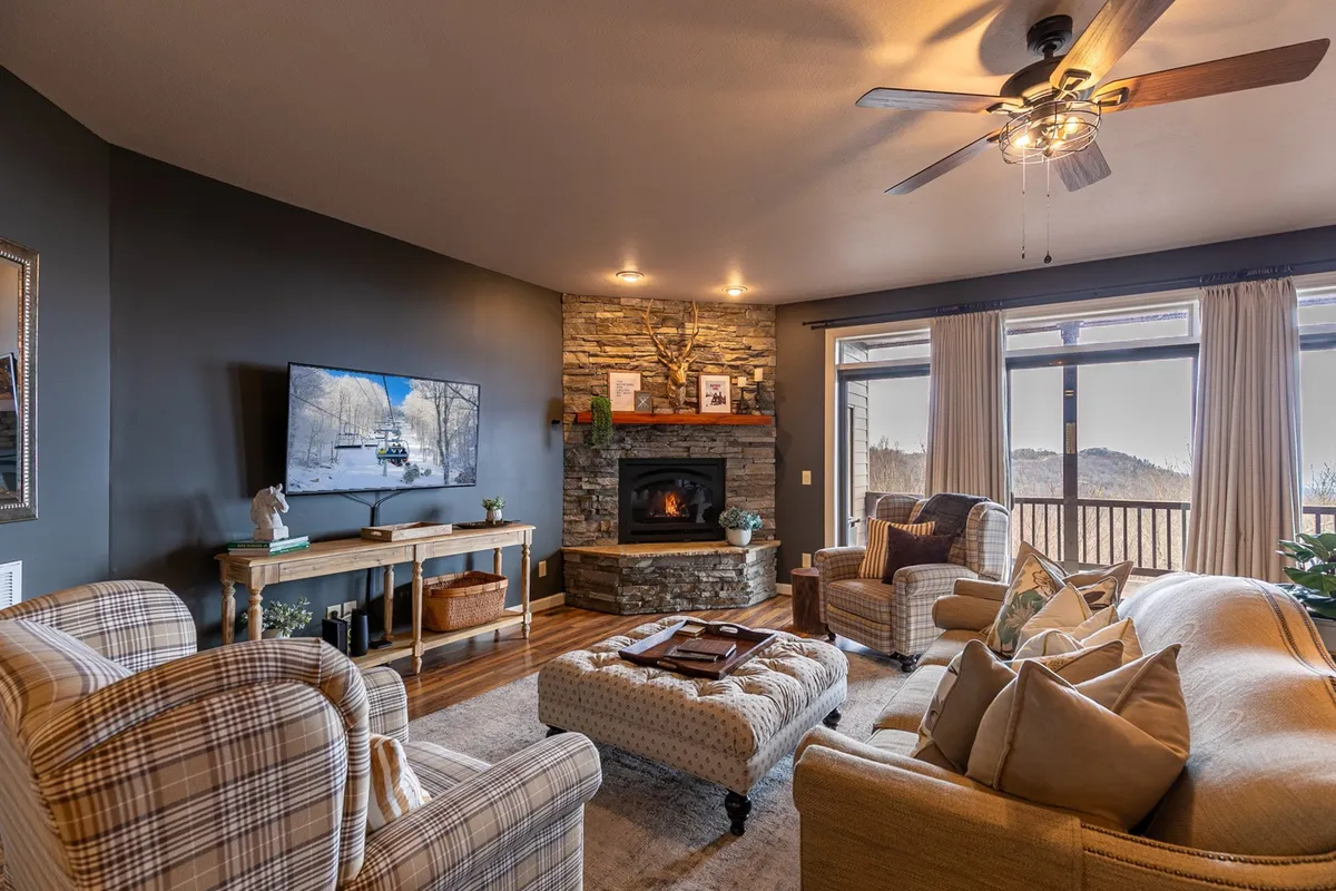The image shows another angle of the living room, focusing on the fireplace, television, and comfortable seating, but also incorporating a window with mountain views. It conveys a sense of warmth and togetherness, with the natural scenery visible, aligning with the section’s emphasis on both indoor coziness and outdoor vistas.