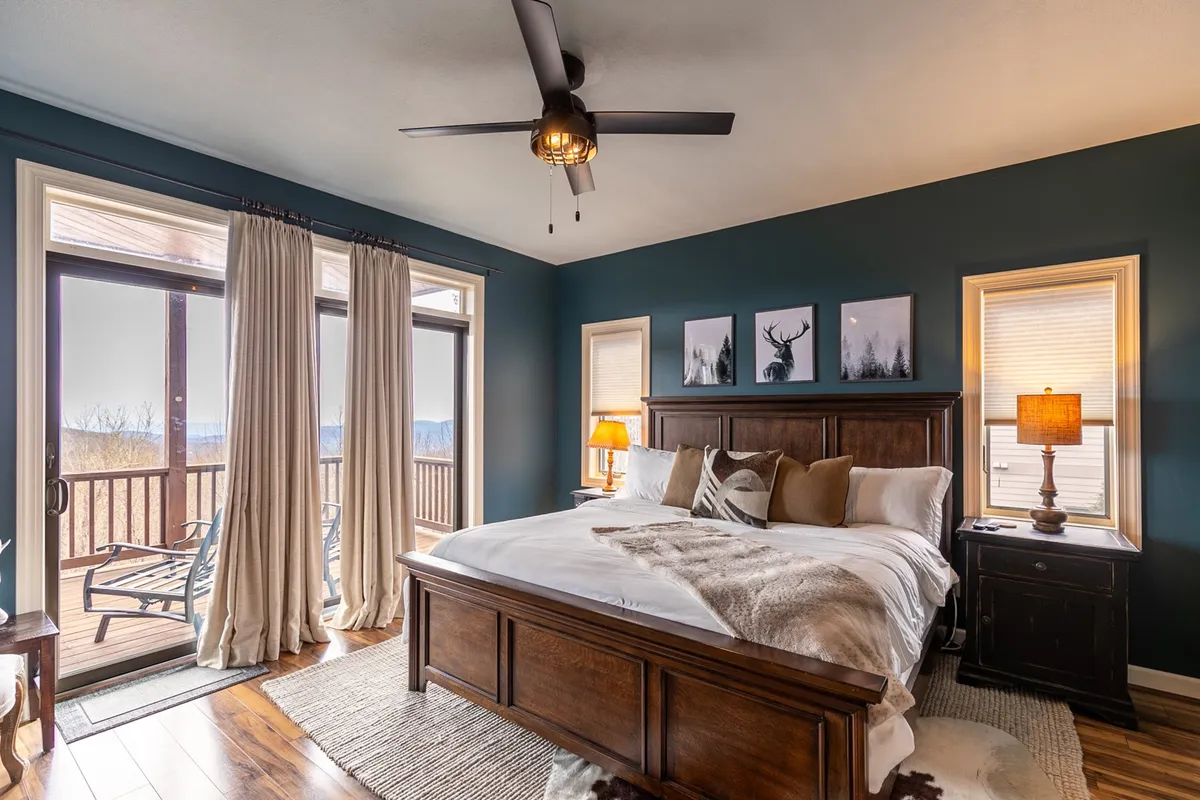 A serene king-sized bed is framed by wooden nightstands and large windows with curtains drawn aside, offering sweeping views of the mountains just beyond. The tranquil setup invites relaxation and provides a direct visual connection to the surrounding landscape.