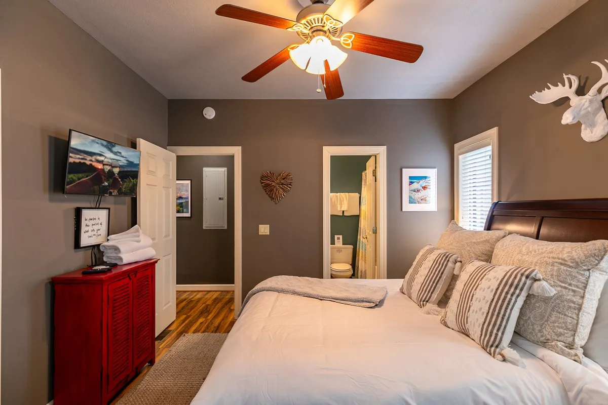 A cozy king-size bedroom featuring a plush bed adorned with decorative pillows, a red wooden dresser, ceiling fan with a light fixture, and tasteful wall art and framed pictures. The room is bathed in natural light, creating a restful and inviting atmosphere.